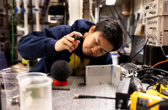 A man sits at a steel workbench with a soft-ball sized sphere resting on a platform placed in front of him. He holds a t-shaped tool a few inches away from the sphere and peers at it from the side.