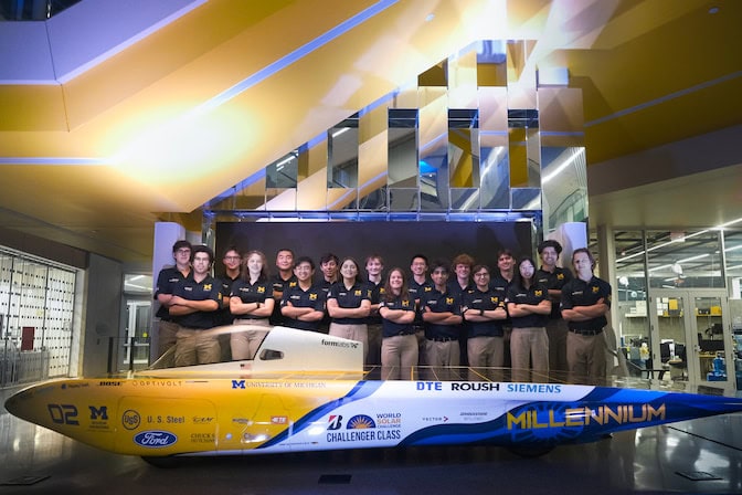 All 18 members of the solar car race crew stand behind Millennium with their arms crossed. Most of the vehicle is roughly knee high and covered in solar cells, with the top of the cockpit at hip height.