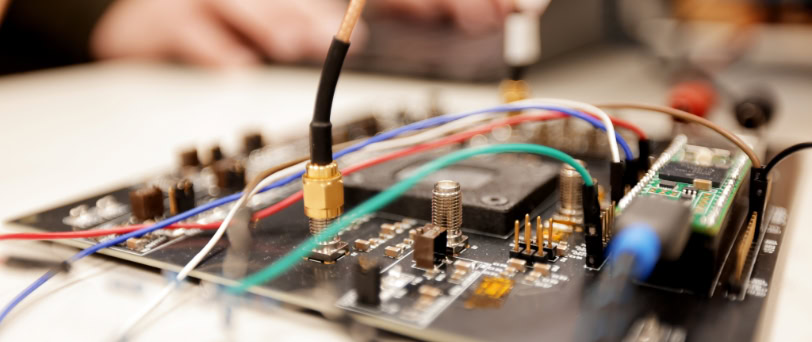 A circuit board with green, blue, red, white and yellow wires extending from it. Gold and black knobs surround a raised black square in the middle. The square holds a computer chip.