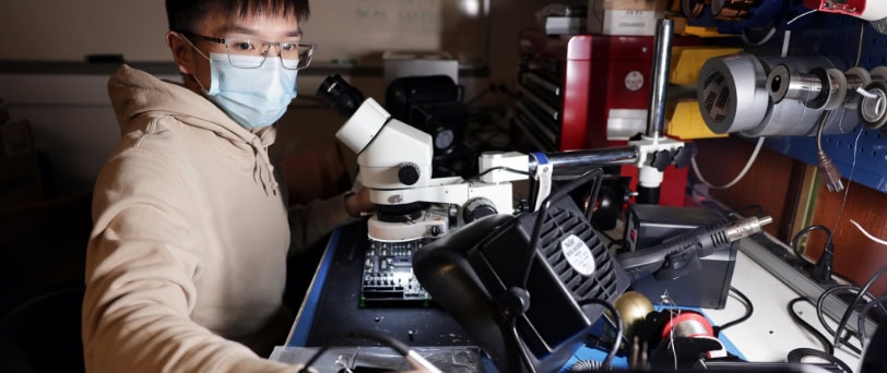 A young man with short dark hair wearing a mask, glasses and a sweatshirt stands at a table full of lab equipment including a microscope. He is writing something down.