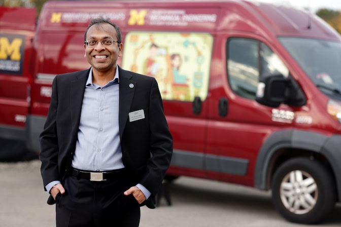 A man in a dark jacket stands with hands in pockets in front of a large red van.