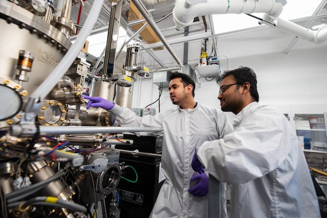 Two men wearing white lab coats and purple nitrile gloves stand together, inspecting a valve on a large silver and gold machine.