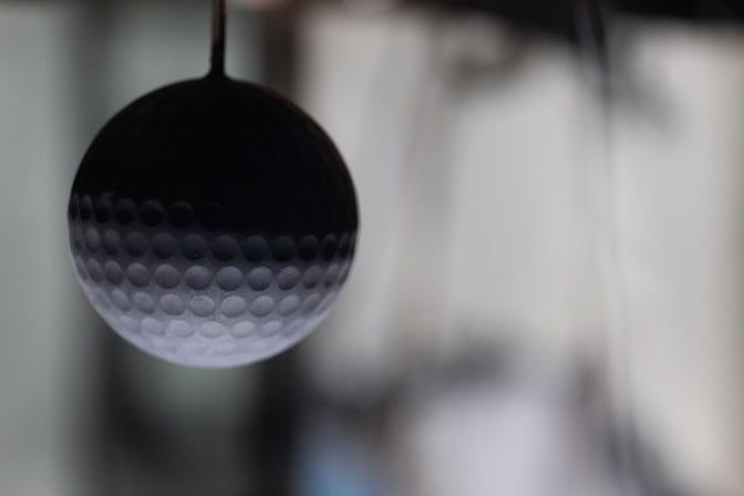 A blurred background with a black, softball-sized sphere with dimples like a golf ball is suspended by a thin rod in the foreground.