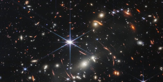 Thousands of small galaxies appear across this view. Their colors vary. Some are shades of orange, while others are white. Most appear as fuzzy ovals, but a few have distinct spiral arms. In front of the galaxies are several foreground stars. Most appear blue, and the bright stars have diffraction spikes, forming an eight-pointed star shape. There are also many thin, long, orange arcs that curve around the center of the image. Please reference the extended text description for more details.