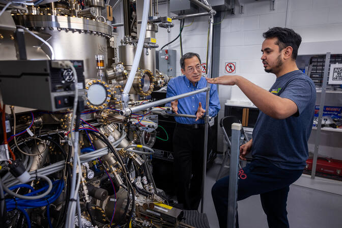Two men stand facing each other in conversation, next to a large silver and gold machine with many tubes and valves branching off of it.