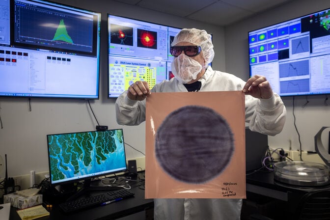 A man in a hair net, beard net, lab coat and protective eyeglasses holds up a piece of paper with a 12-inch gray circular mark in its center. The gray is slightly deeper in some areas and lighter in others, as if colored in pencil over wood grain. Behind, screens show graphs visualizing characteristics of the beam.