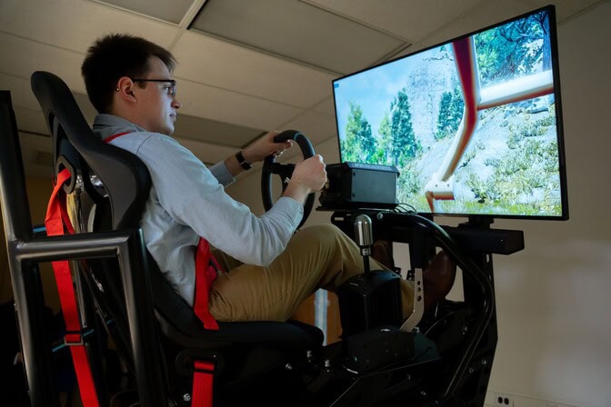  A man is securely strapped into a motion-based driving simulator seat, hands on a steering wheel and eyes on a screen a few feet ahead, mounted on the moving base. The screen displays rocky terrain with tufts of grass and conifers as well as the part of the roll cage that would be in his line of sight in the virtual vehicle he is testing.