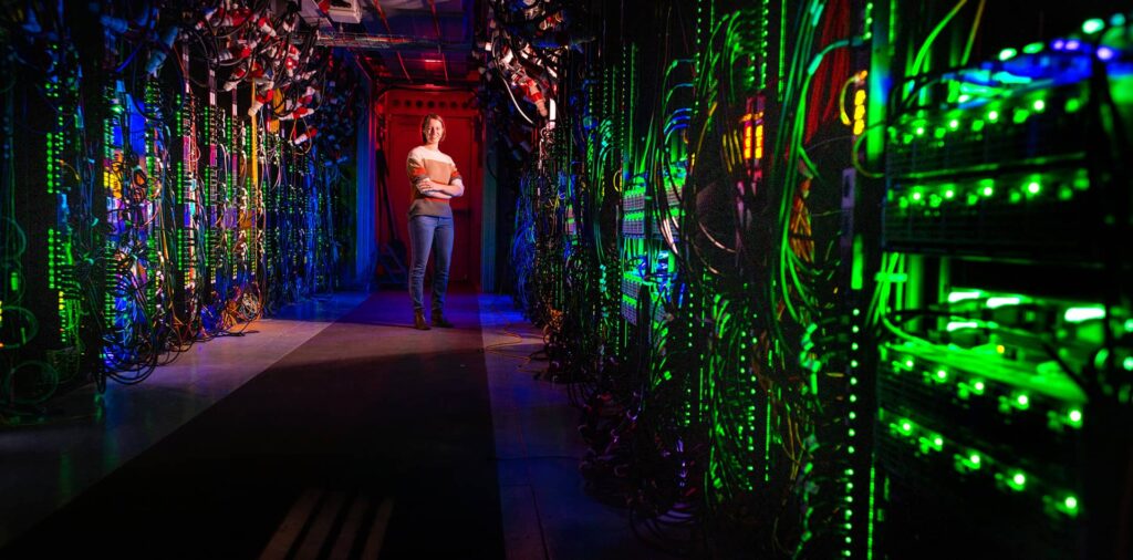 A woman stands inside a high-tech data center, surrounded by rows of computer racks and ambient lighting.