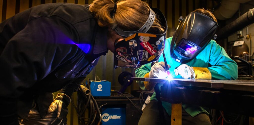 Two people in protective gear perform welding indoors.