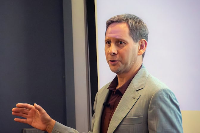 Medium shot of a man wearing a gray blazer and burgundy golf shirt speaking and gesturing with his hand.