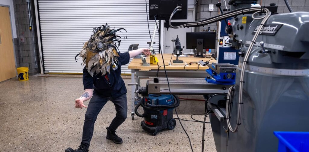 A woman wearing a face mask poses for a photo in a well-lit lab filled with various fabrication tools and equipment.
