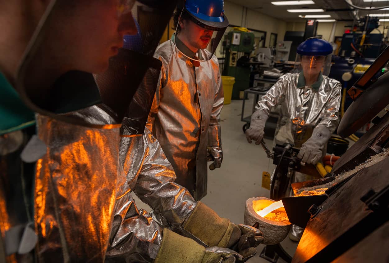A group of people wearing safety gear pour molten bronze into molds in a workshop setting, casting tools they designed in a university engineering class.