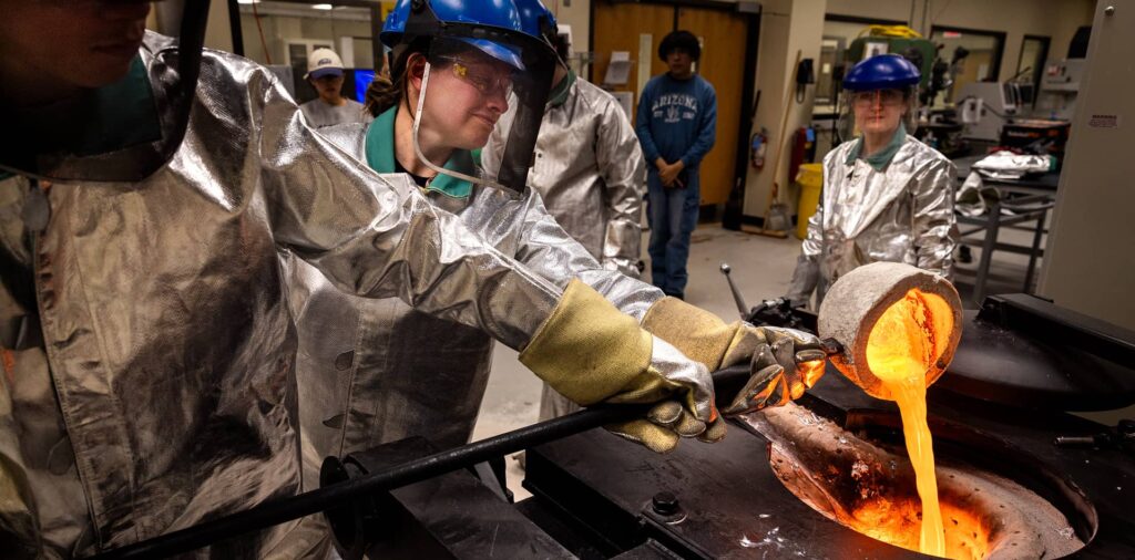A group of people wearing safety gear pour molten bronze into molds in a workshop setting, casting tools they designed in a university engineering class.