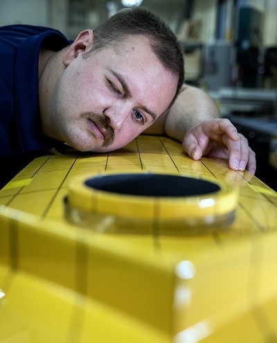 A man closes his right eye as he leans over the smooth, yellow surface of a model ship.