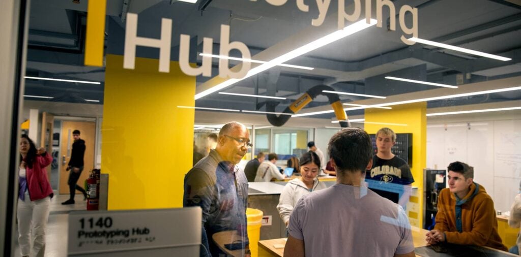 A group of students and a staff member discuss a medical equipment prototype in a university lab setting.
