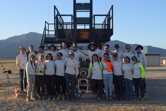 21 students in matching team shirts flank the engine, which is shown end-on—a circular metal structure about 8 inches wide. The ground is sandy, with mountains in the distance.