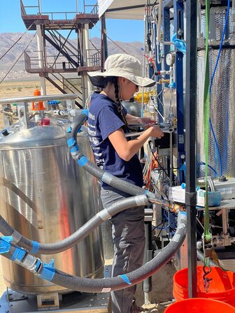 A student uses a wrench to tighten a valve, standing in the middle of a complex structure of tanks and pipes.