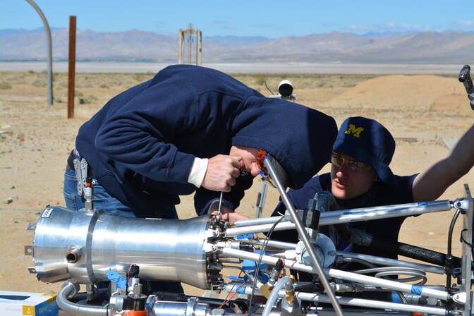 With the Mojave Desert mountains in the background, two students from the MASA hotfire team make adjustments to their engine—a cylindrical metal structure about 8 inches wide connected on one end to pipes, sensors, and wiring.