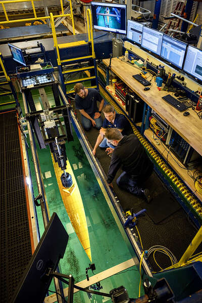 Three engineers kneel on the lower platform of a steel carriage. They are leaning over a channel cut into the center of the platform to inspect the ship model, which is lowered into the underlying water through the channel. The engineers are surrounded by an assortment of electrical and mechanical equipment, as well as computer monitors displaying data in graphs and spreadsheets.