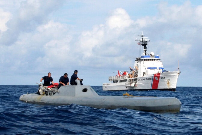 Three members of the coast guard stand on the deck of a grey vessel. The deck sits only a few feet above the water. A red and white Coast Guard ship is anchored behind the grey vessel.