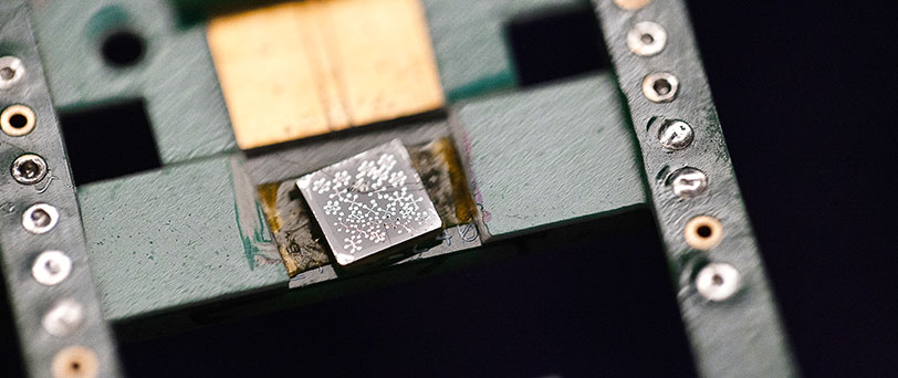 A small silver chip with patterns that resemble flowers on its surface is mounted on a green platform with various circular metal contacts.