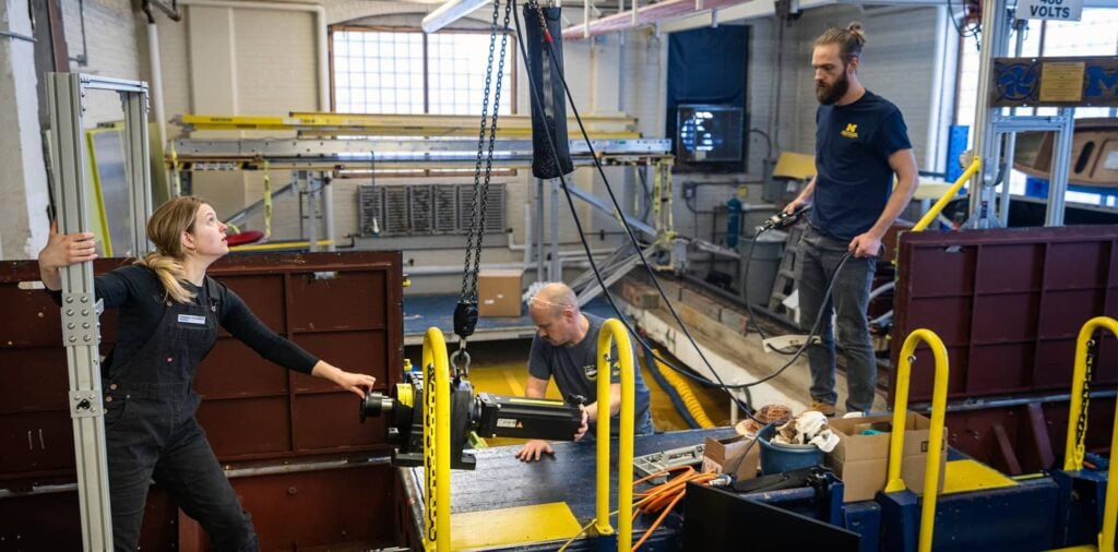 Three individuals work together to install a new motor onto a large mechanical carriage in a research lab, surrounded by engineering equipment and tools.