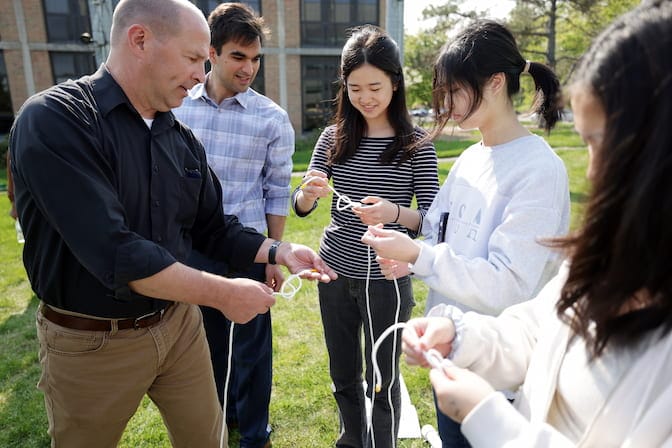 Five people—two adult men on the left and three young women on the right—stand in a grassy field in front of a high school building. One man demonstrates how to tie a knot in a guyline rope. The three young women are tying the same knot with their own ropes.