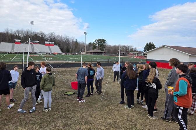 A group of about 20 high school students stand in a grassy area in front of a football field. They are gathered near two sets of PVC pipe poles connected by a wire with guyline ropes tethering them to the ground. Recently used tools—a bucket, clip board, and wires—lie on the ground between the PVC pipe poles.