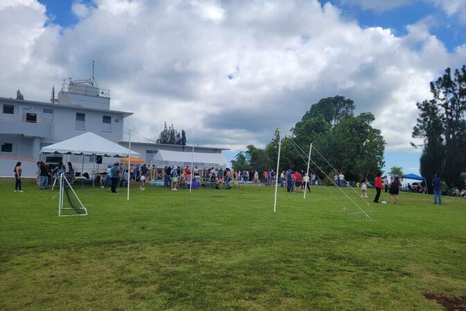 Foreground: Two sets of 10 foot tall PVC pipe poles connected by a wire with guyline ropes tethering them to the ground stand in a grassy area with a small soccer goal to the left. Background: Two tents are set up outside a building. People stand under the tent at tables or stand in groups.