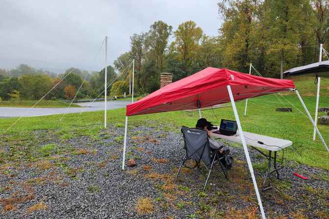 Foreground: A red tent stands over a white card table in a gravel patch surrounded by grass. A teenage boy sits at the table in a lawn chair looking a laptop in front of him. In the grass behind the red tent stands two sets of 10 foot tall PVC pipe poles connected by a wire with guyline ropes tethering them to the ground.