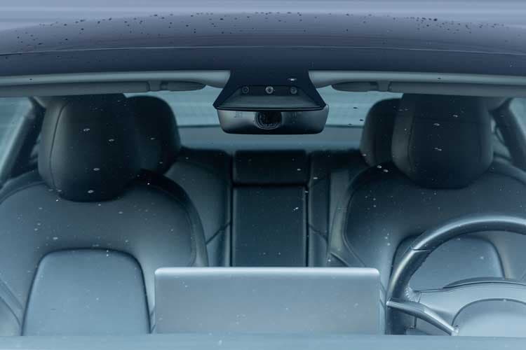 A vehicle's cabin through its front windshield, showing two empty seats and a steering wheel. A small black box is in the top center of the vehicle interior.