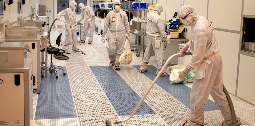 Individuals in full protective gear stand in a cleanroom lab, cleaning and doing maintenance tasks, while holding cleaning equipment and trash bag.