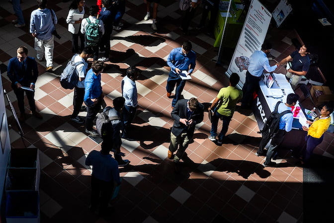 Viewed from overhead, college students dressed in business clothes walk through an atrium. Sunlight streams in low, casting long shadows.