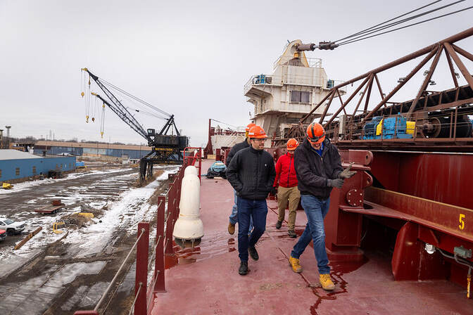 People wearing orange hard helmets walk alongside a commercial ship docked at a port.