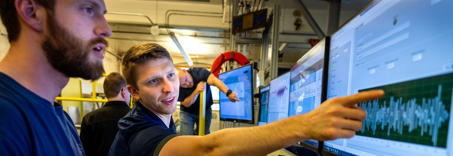 Two engineers stand in front of several computer monitors on the tow tank carriage. One engineer is pointing at data on a graph, which looks like a complex, oscillating wave pattern.