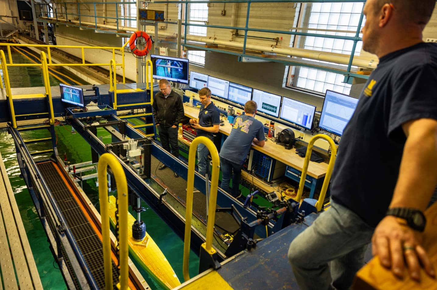 A man watches a team of engineers from an elevated platform. The engineers are working on a lower platform near a ship model. The slender, yellow model is lowered into the water through a channel cut into the lower platform. A steel mast connects the model to rails on the elevated platform.