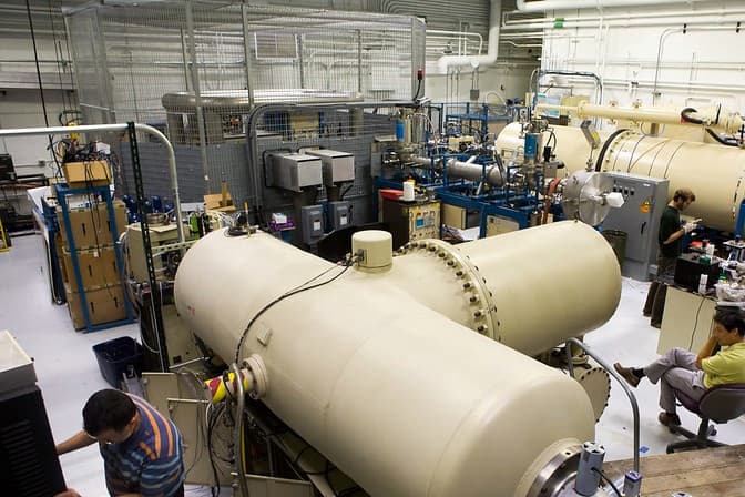 A high-tech laboratory with three researchers working among large cylindrical machines, control panels, and various lab equipment.