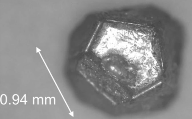 A silvery, metallic quasicrystal grain resembles a 12-sided die used in tabletop games. A scale bar shows the diameter of the grain is 0.94 millimeters.