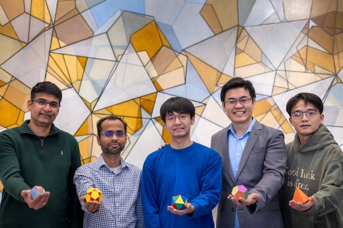 Five men stand in front of a wall, decorated with overlapping orange, gray and blue shapes. Each man holds a large die-like object. From left to right, the men hold: a crystal with rhombus-shaped faces; a crystal with pentagonal and triangular faces; a crystal with 20 triangular faces; a crystal with 12 pentagonal faces, and a four-faced pyramid.