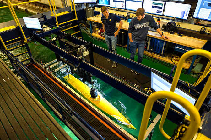 Two researchers stand on a platform above a vessel while they conduct resistance and seakeeping tests of a Low-Profile Vessel at the Marine Hydrodynamics Laboratory.