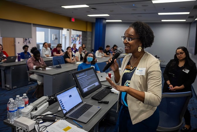 A medium shot of a woman standing in front of a room full of people, speaking into a microphone and gesturing with her hands. She wears a beige blazer, colorful shirt and gold earrings.