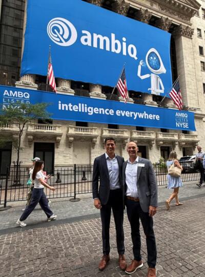 Two men wearing suits stand on a brick street in front of an ornate building. A large blue banner behind them says "ambiq." A narrow banner beneath it says "intelligence everywhere." Behind them, people walk on the sidewalk.