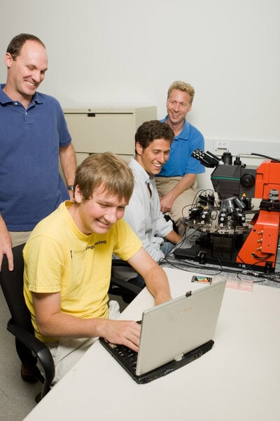 Fojtik works at a computer while Hanson sits at a device that looks like a complicated microscope. Sylvester stands behind Fojtik and Blaaw sits behind Hanson. All four men are smiling in the general direction of Fojtik's computer.
