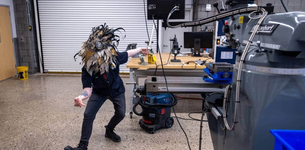 A woman wearing a face mask poses for a photo in a well-lit lab filled with various fabrication tools and equipment.