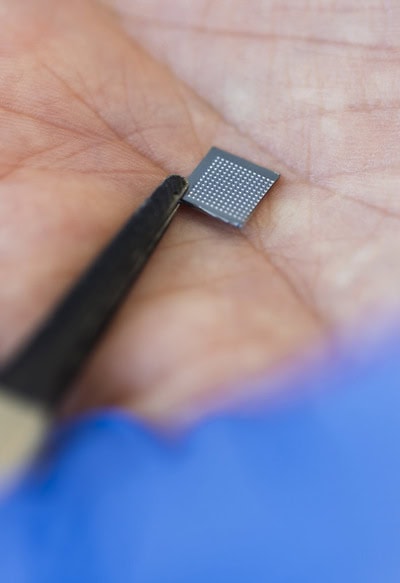 A close up of a palm holding a square silicone patch, just a few millimeters wide, that has tiny microneedles on one side. Tweezers hold the patch in place.