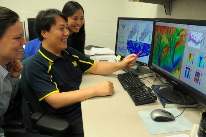Three people discussing visualizations on dual computer monitors.