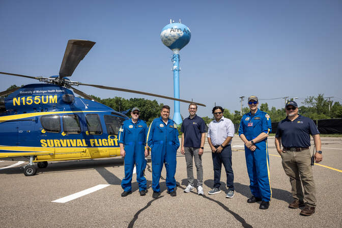 The helicopter is to the left, painted in blue and yellow with "SURVIVAL FLIGHT" on the side. Newell, Adelmann and Vanderberg are in flight suits, McGuire and Lollar wear Mcity polos, and Viswanathan wears a button-down shirt.