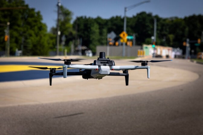 A black and white quadcopter, about the size of a board game box, hovers over a roundabout.