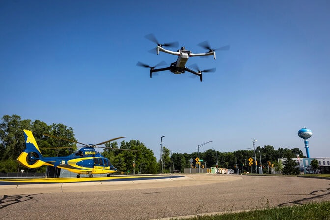 A black and white quadcopter, about the size of a board game box, hovers over a roundabout. A blue and yellow helicopter is parked in the background.