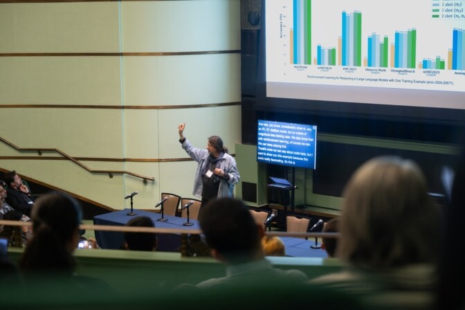 A man stands with his arm upraised, a bar chart displayed on the large screen behind him, and auto transcription on a smaller screen.
